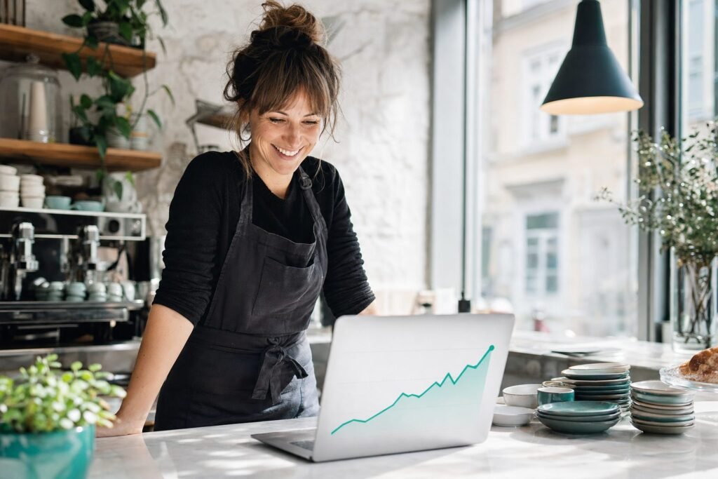 Smiling café owner at her counter checking a laptop that shows a rising teal growth chart